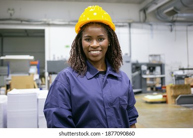 Cheerful African American Female Factory Employee In Hardhat And Overall Standing On Plant Floor, Looking At Camera And Smiling. Front View, Medium Shot. Women In Industry Concept