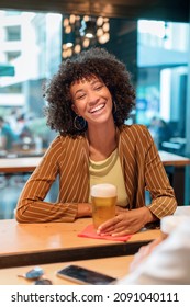 Cheerful African American female with short curly hair in casual clothes sitting at wooden bar counter with glass of beer and laughing in pub in daytime