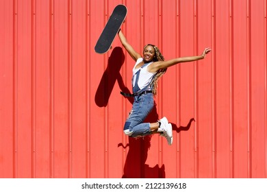 Cheerful African American Female Skater With Afro Braids And Skateboard Jumping Above Ground Against Red Wall In City And Looking At Camera