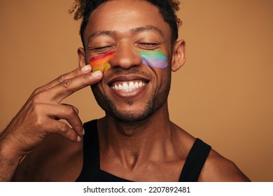 Cheerful African American gay smiling with closed eyes and painting rainbow on cheeks during LGBT pride event against brown background - Shutterstock ID 2207892481