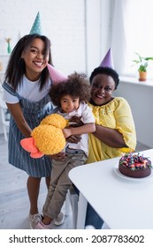 Cheerful African American Girl Holding Soft Toy While Mom And Granny Hugging Her Near Birthday Cake