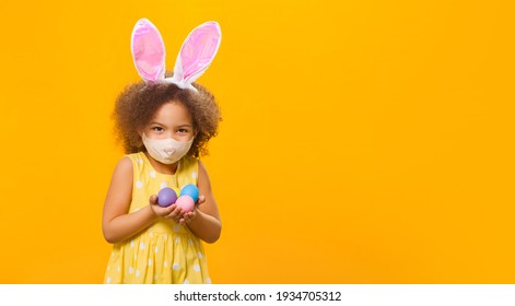 A Cheerful African American Girl With Rabbit Ears On Her Head And A Protective Mask With A Few Colored Eggs In Her Hands On A Yellow Background. Funny Happy Baby. Covid Easter To The Child.