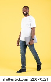 Cheerful African American Guy Holding Laptop Computer Posing Walking Over Yellow Studio Background. Freelance Career And Internet Business Concept. Vertical, Full Length Shot