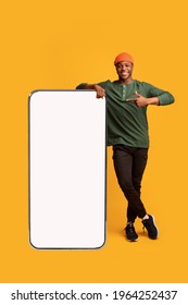 Cheerful African American Guy Leaning And Pointing At Big Smartphone With White Blank Screen, Showing Free Copy Space For Your Mobile App Or Website Design, Standing On Yellow Background, Mockup Image