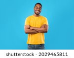 Cheerful African American Guy Smiling To Camera Posing Crossing Hands Standing Over Blue Background. Studio Shot Of Happy Self-Confident Black Millennial Man Expressing Positive Emotions