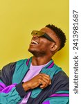 cheerful african american guy in trendy sunglasses and bright jacket laughing on yellow, streetwear