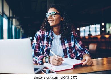 Cheerful African American Hipster Girl In Eyewear Writing Homework In Notepad While Looking Away Studying At Laptop.Positive Teen With Curly Hair Thinking On Plans Sitting With Notebook At Desktop