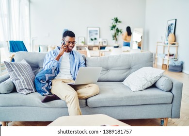 Cheerful African American Hipster Guy Having Mobile Phone Talk While Working Remotely From Home On Netbook, Smiling Male Freelancer Calling To Banking Service Making Transaction On Laptop Computer
