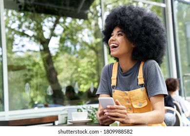 Cheerful African American hipster woman using phone, laughing, sitting at outdoor cafe table. Smiling happy mixed race black lady with afro hair having fun or feeling excited holding smart phone.