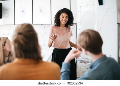 Cheerful African American Lady With Dark Curly Hair Standing Near Board And Happily Looking At Her Colleagues In Office. Young Beautiful Business Woman Giving Presentation To Coworkers During Meeting