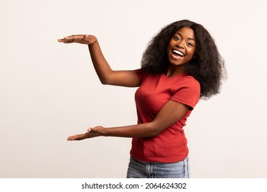 Cheerful African American Lady Holding Invisible Object With Her Hands, Happy Excited Black Woman Measuring Something With Arms While Standing On White Background, Recommending New Item, Copy Space