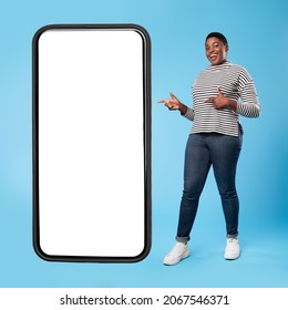 Cheerful African American Lady Standing Near Big Smartphone With Blank Screen And Pointing Fingers Advertising Mobile Application Standing Over Blue Studio Background. Square Shot, Mockup