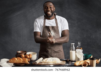 Cheerful African American Male Cook Presents Culinary Talents, Participates In Television Cooking Show, Wears Apron, Stands At Kitchen Table, Makes Dough For Preparing Buns Isolated On Chalk Wall