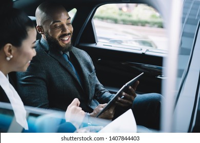 Cheerful African American male director communicating with female secretary during car drive to business meeting discussing startup ideas and reading notes for presentation from digital tablet