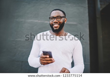Cheerful african american male in spectacles using mobile phone outdoors looking waya and smiling, positive dark skinned hipster guy in white shirt with copy space for brandname holding cellular