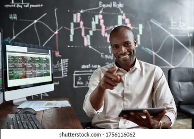 Cheerful african american male trader sitting by desk, pointing at camera and studying analytical reports using tablet pc in the office. Stock trading, people concept. Horizontal shot. Front view