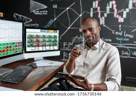 Cheerful african american male trader sitting by desk, pointing at camera and studying analytical reports using tablet pc in the office. Stock trading, people concept. Horizontal shot.