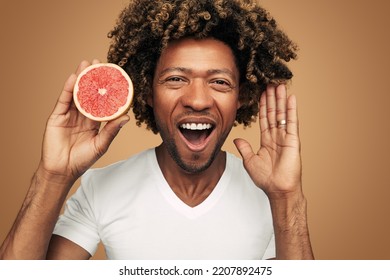 Cheerful African American Male In White T Shirt With Curly Hair Raising Arm And Looking At Camera, With Opened Mouth While Demonstrating Half Of Fresh Grapefruit Against Brown Background