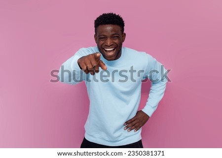 cheerful african american man in blue sweater taunts and sneers points his hand at the camera on pink isolated background, the man jokes and makes fun of you and laughs