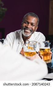 cheerful african american man clinking beer glasses with blurred senior friends in pub