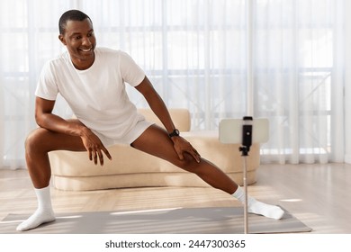 A cheerful African American man is doing a side lunging stretch in front of a smartphone mounted on a tripod, likely filming a workout or instructional video - Powered by Shutterstock