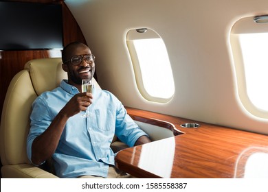 Cheerful African American Man In Glasses Holding Champagne Glass In Private Plane 