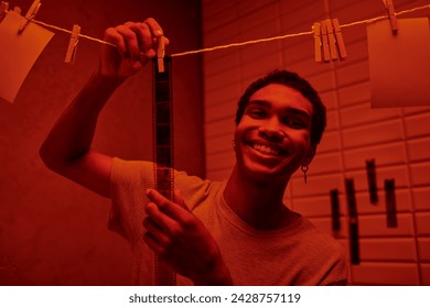 cheerful african american man hanging freshly developed film strip  in a red-lit darkroom, nostalgia - Powered by Shutterstock