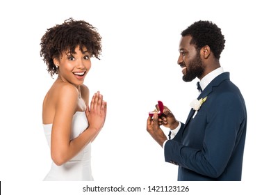 cheerful african american man holding box with ring while making proposal to surprised woman isolated on white 