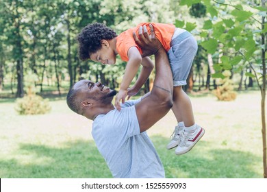 Cheerful African American Man Holding Son Above Head While Walking In Park