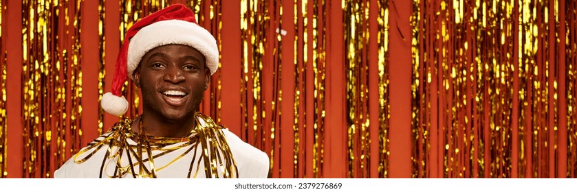 cheerful african american man in santa cap smiling at camera near shiny tinsel on red, banner - Powered by Shutterstock