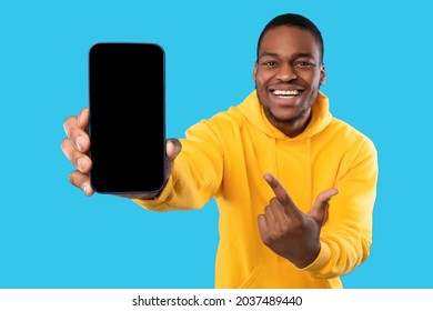 Cheerful African American Man Showing Cellphone With Blank Screen Advertising Great Application And Pointing Finger At Smartphone Posing Over Blue Studio Background. Check This New App. Mockup
