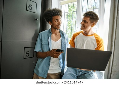 cheerful african american man with smartphone talking to student using laptop near locker in hostel - Powered by Shutterstock