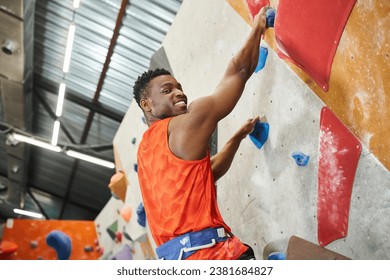 cheerful african american man smiling happily at camera while climbing up rock wall, bouldering - Powered by Shutterstock