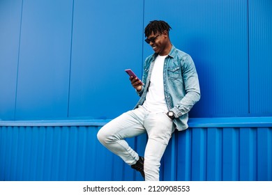 Cheerful african american man in stylish clothes and sunglasses browsing social media photos on mobile phone enjoying free time outdoors, happy black man standing against blue wall of Shopping Mall
