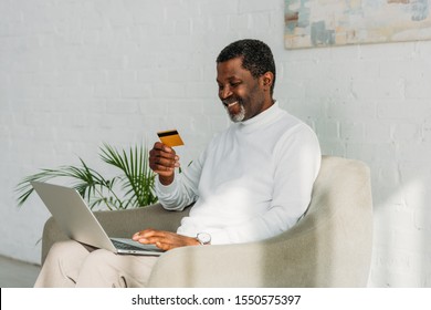 Cheerful African American Man Using Laptop And Holding Credit Card While Sitting On Sofa