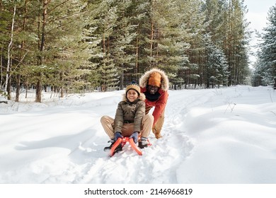 Cheerful African American man in winterwear pushing sledge with his son while moving along road covered with snow in forest or park