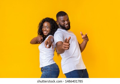 Cheerful African American Man And Woman Pointing Fingers At Camera, staying back to back on yellow studio background, free space