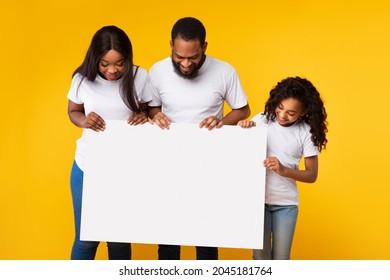 Cheerful African American man, woman and girl looking down at empty advertising placard, yellow studio background. Smiling black family holding blank board for advertisement or text in hands - Powered by Shutterstock