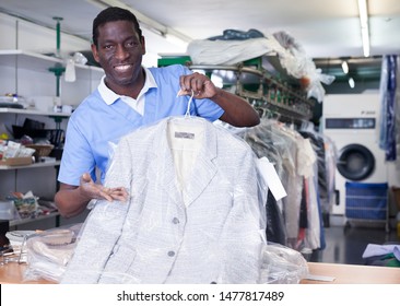 Cheerful African American Man Worker Of Dry Cleaner Standing At Reception Counter, Showing Clean Clothes After Dry Cleaning