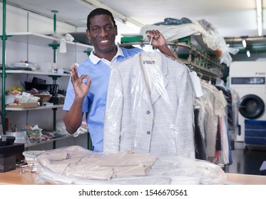 Cheerful African American Man Worker Of Dry Cleaner Standing At Reception Counter, Showing Clean Clothes After Dry Cleaning