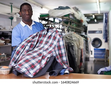 Cheerful African American Man Worker Of Dry Cleaner Standing At Reception Counter, Showing Clean Clothes After Dry Cleaning