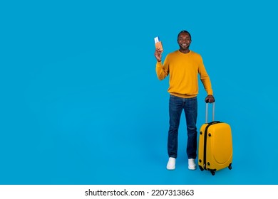 Cheerful African American Middle Aged Man Tourist Travelling Alone, Carrying Yellow Luggage, Showing Passport With Flight Tickets, Posing Over Blue Studio Background, Copy Space, Full Length