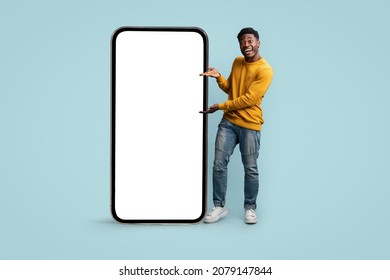 Cheerful african american millennial man showing huge modern cellphone with blank screen, showing newest entertaining or dating mobile app, mockup, blue studio background, copy space