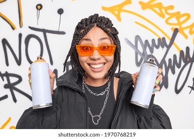 Cheerful African American Millennial Woman With Braids Holds Two Aerosol Spray Bottles Being Creative Street Artist Draws Graffiti Wears Fashionable Clothes Orange Sunglasses Belongs To Gang. Swag