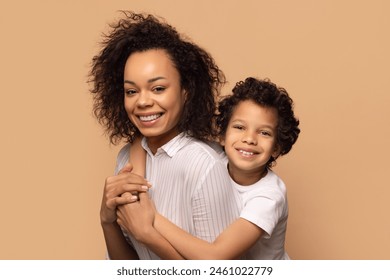 A cheerful African American mother with curly hair is holding her young son lovingly as they both smile broadly. They are posing against a neutral beige background, closeup - Powered by Shutterstock