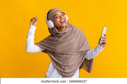 Cheerful african american muslim girl holding smartphone, listening music in wireless headphones and dancing over yellow background with free space - Powered by Shutterstock