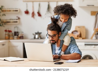 Cheerful African American Playful Kid Sitting On Neck Of Unhappy Busy Dad During Working With Laptop In Home Kitchen