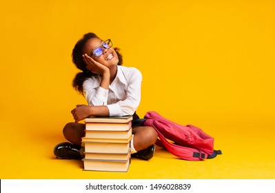 Cheerful African American Schoolgirl Dreaming Sitting At Book Stack Over Yellow Background In Studio. Copy Space - Powered by Shutterstock