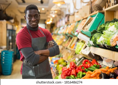 Cheerful african american seller standing with crossed arms near counter with fresh fruits and vegetables in store..