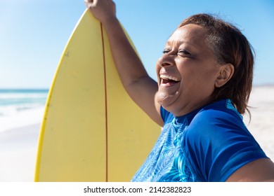 Cheerful african american senior woman standing by yellow surfboard at beach on sunny day. unaltered, active lifestyle, aquatic sport and holiday concept. - Powered by Shutterstock